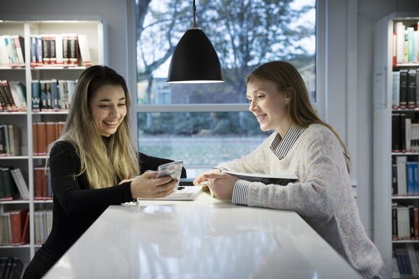 Two students looking at a mobile phone at a library. Photo.