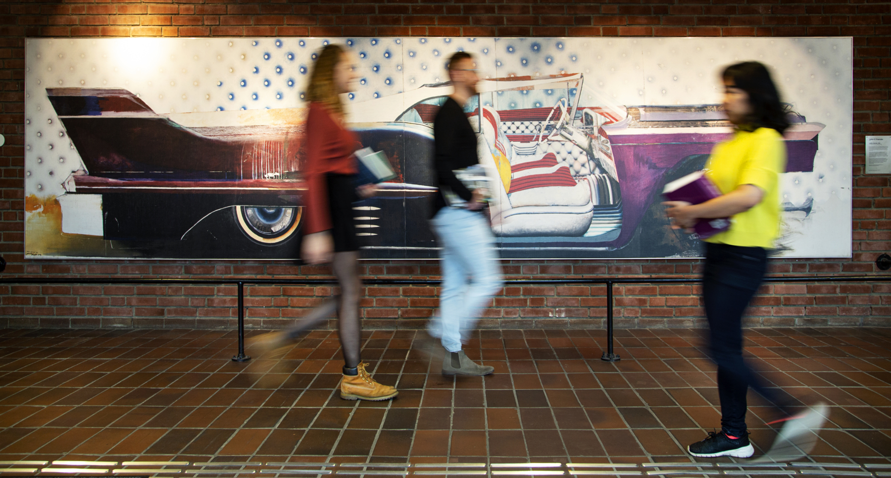 Tre studenter i suddiga konturer går framför konstverket Cadillac Eldorado i V-huset på LTH. Foto.