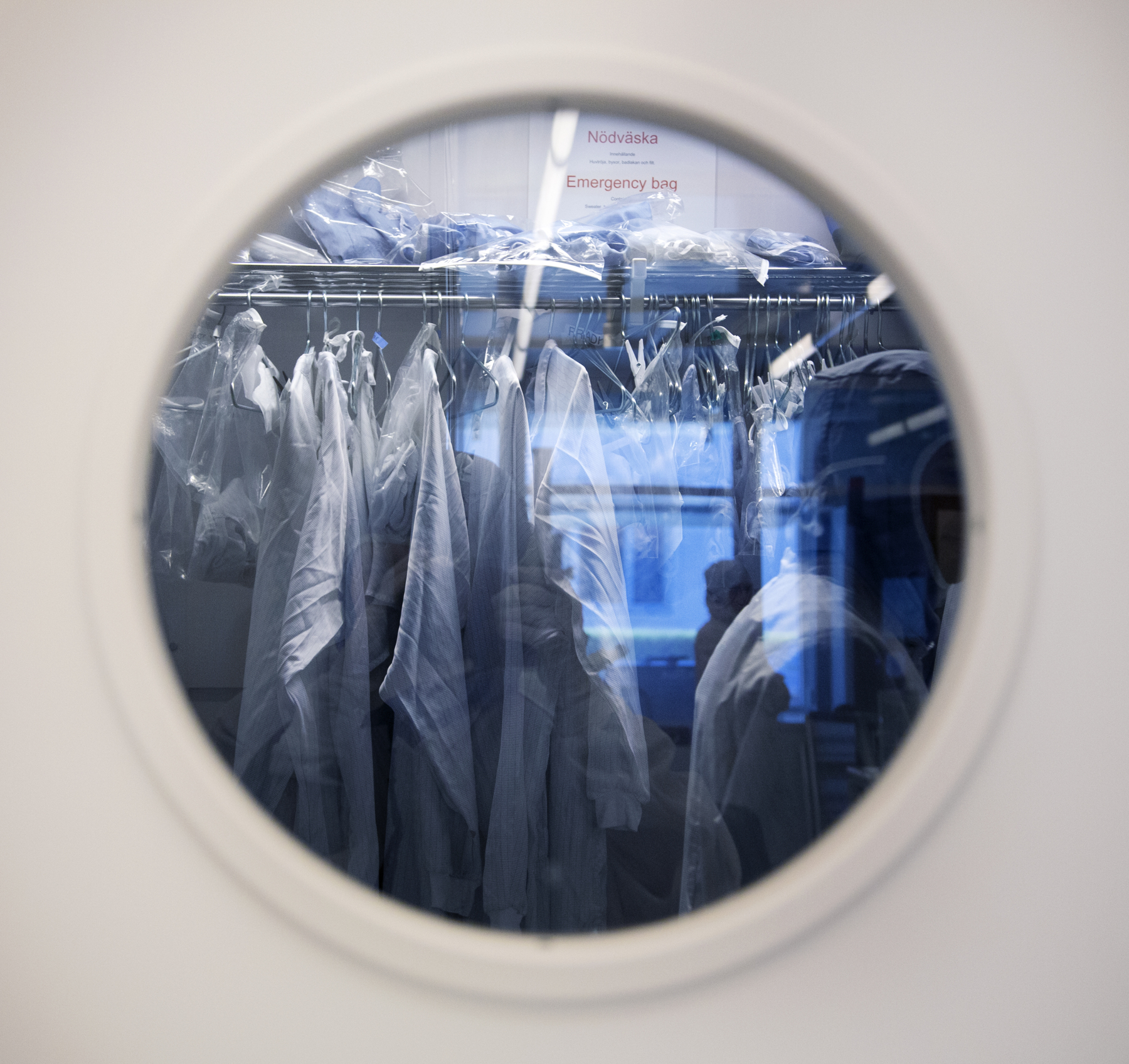 Genom runt fönster i vit dörr syns labbrockar hänga, i ett blått ljus. Foto.