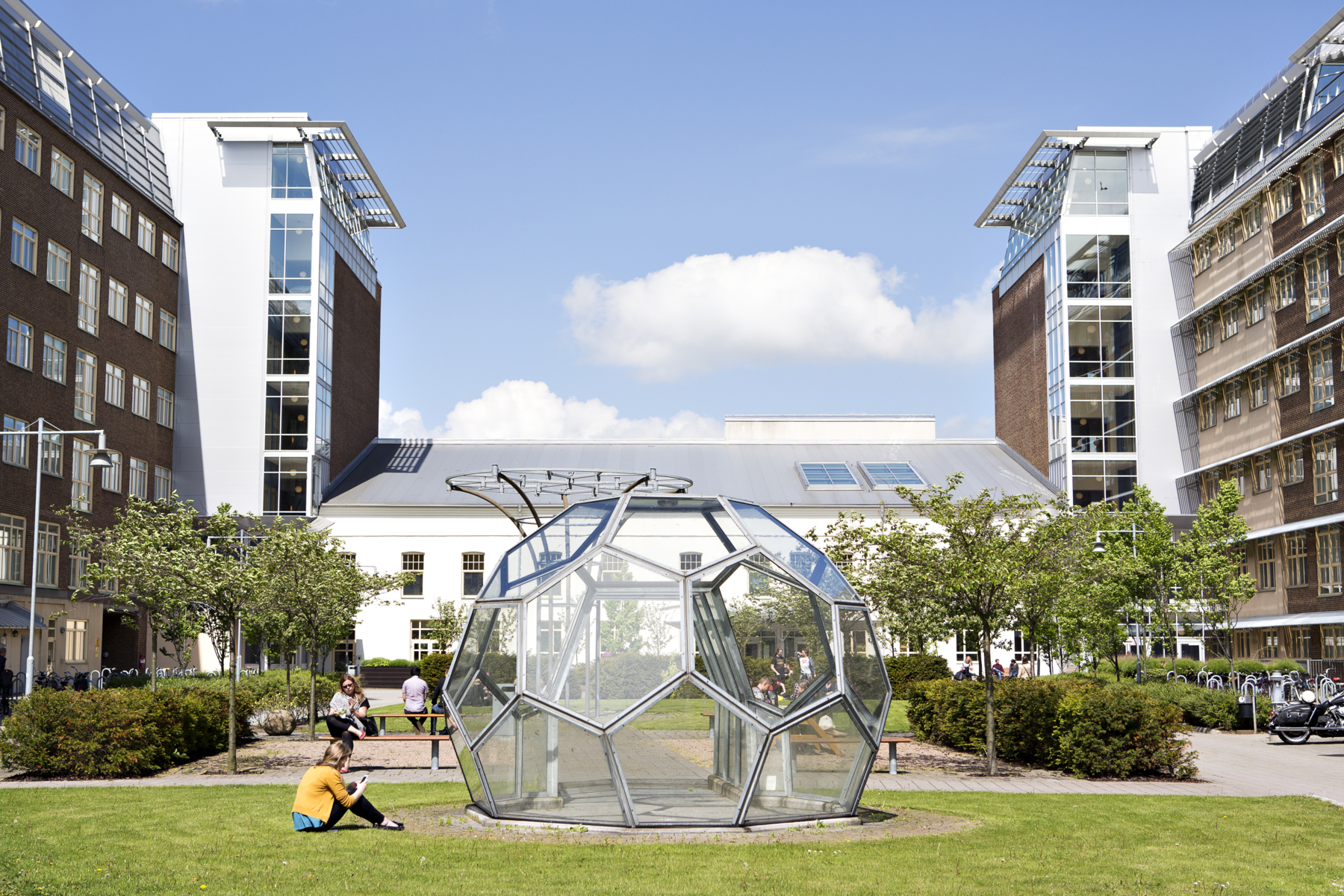 Photo of Campus Helsingborg on a sunny day. A person is sitting in the grass in front of a big glass building.