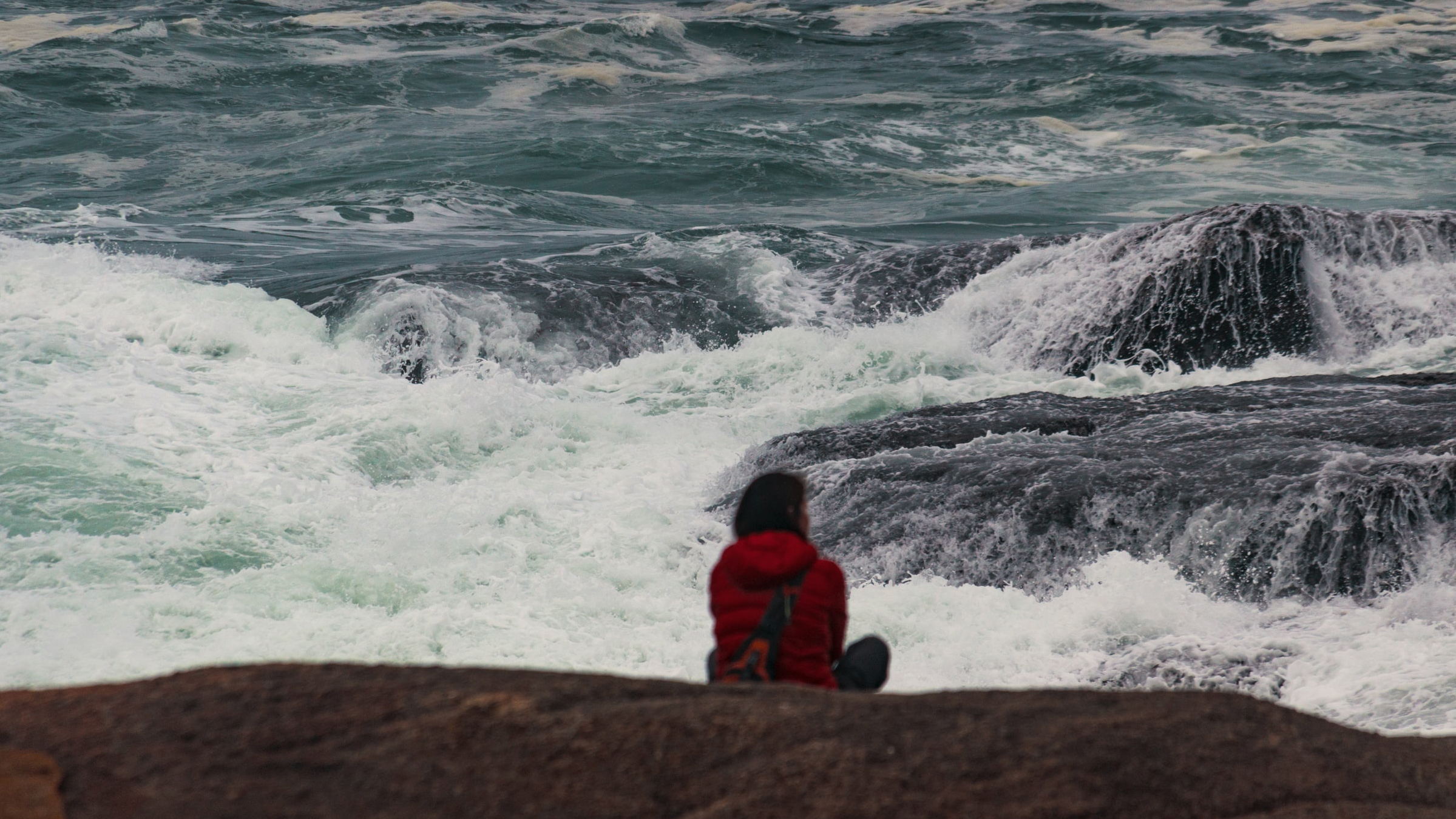 Photo of woman sitting on rocks looking at a stormy sea.