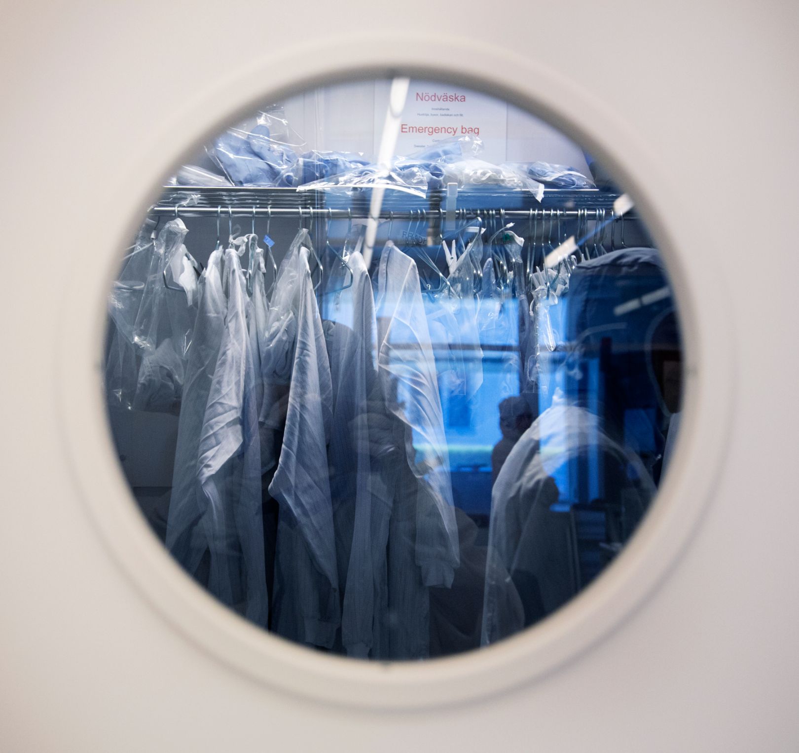 Runt fönster i en vit dörr, bakom vilket vita labbrockar syns. Foto av Charlotte Carlberg Bärg.
