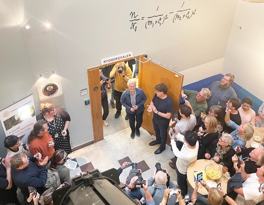 Anne L'Huillier entering a room filled with press and other guests.