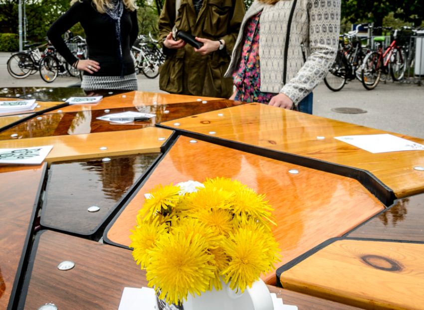 Common dandelions on a wooden table outdoors. three unidentifiable women seem to interact behind it.