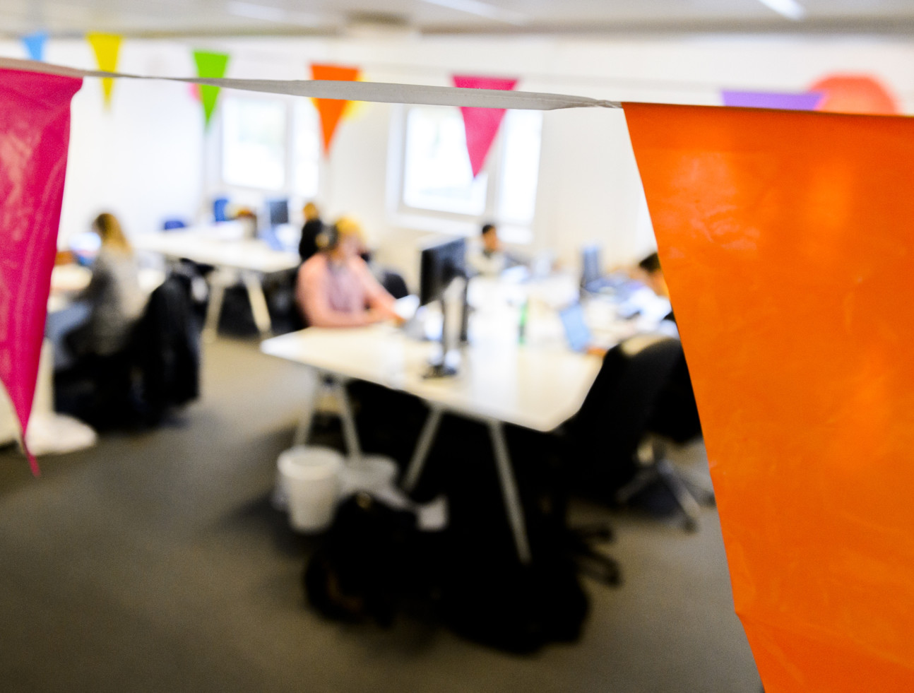 Personer arbetar i ett kontorslandskap, de syns bakom färgglada vimplar i taket. Foto.