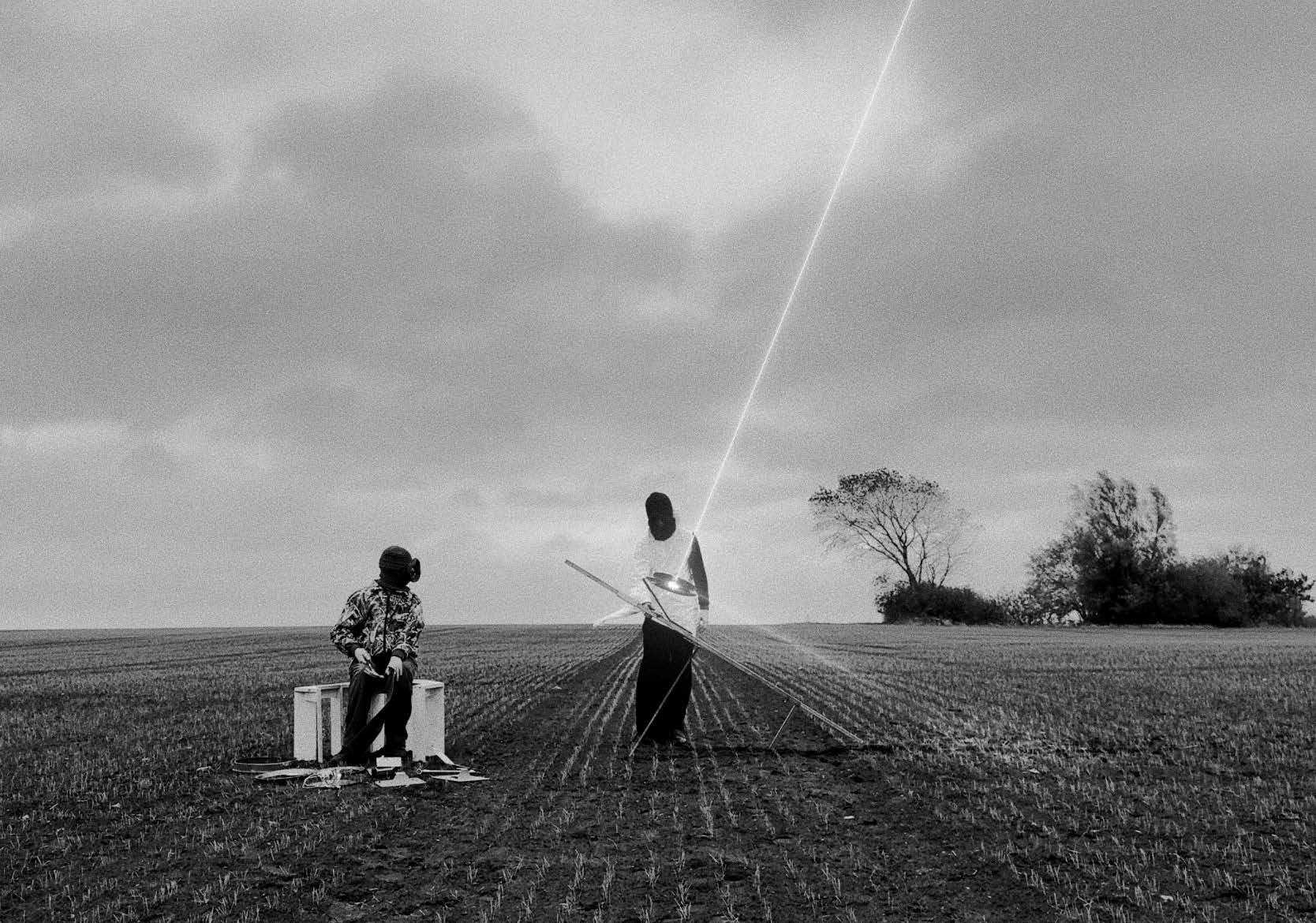 Två maskerade personer står i en åker och från ett föremål lyser vad som ser ut som en laserstråle.