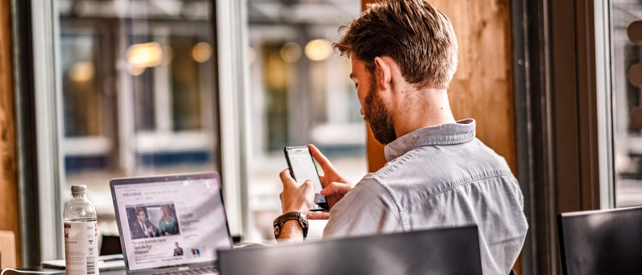 Person sitting behind a laptop, looking att mobile phone. photo.